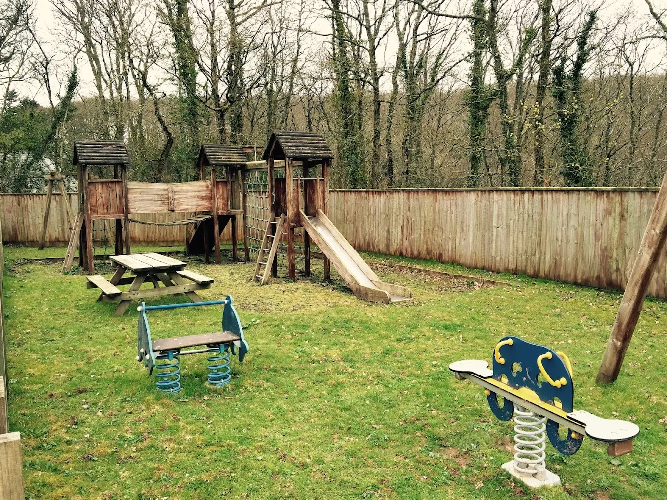 Outdoor wooden play area beside the hall — climber with platform, slide, swings, and spring-mounted ride-on toys on a grassed enclosure